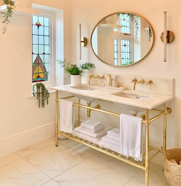 Phòng tắm sạch sẽ với bồn rửa mặt đá marble, vòi nước mạ vàng, gương tròn, cửa sổ kính màu và khăn trắng được xếp ngăn nắp.