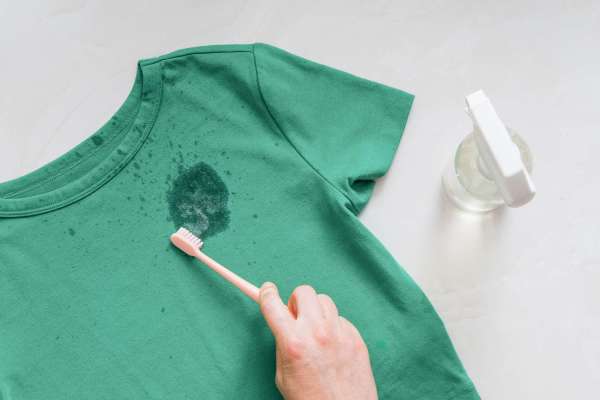 Áo phông màu xanh lá cây bị vấy bẩn đang được chải rửa bởi bàn chải đánh răng, bên cạnh có bình xịt.